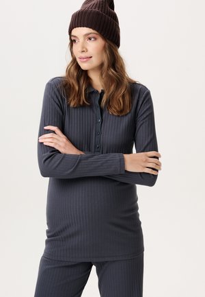 Vrouw met golvend bruin haar die een bruine gebreide muts draagt, een donker geribd shirt met lange mouwen en knopen en bijpassende broek, met gekruiste armen, kijkend naar links.