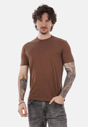 Uomo con capelli ricci che indossa una maglietta marrone e jeans grigi, con braccia tatuate e una posa rilassata con la mano in tasca.