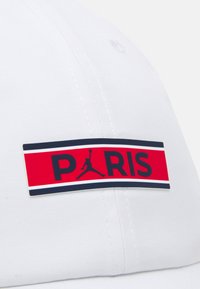 Balta kepurė su priekinės gumos dėklu, kuriame yra užrašas "PARIS" juodu tekstu ant raudono fono, akcentuota jūrine riba ir juodu silueto logotipu.