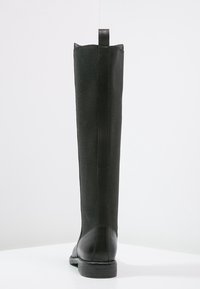 Bota alta de cuero negro con parte superior de tela y tira de agarre, vista desde la parte trasera sobre una superficie y fondo blancos.