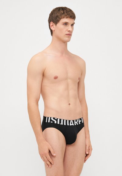 Black brief underwear with a wide waistband featuring bold white lettering. The fabric appears smooth with a snug fit. Model shown from the side.