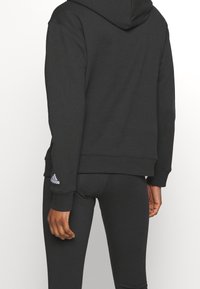 Czarna bluza z kapturem wykonana z miękkiego materiału, z kieszenią kangurka i ściągaczami na rękawach. Logo Adidas w kolorze fioletowym na lewym rękawie.