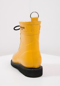 Bota de tobillo de goma amarilla con un acabado texturizado, que presenta una suela negra, un lazo de tirón en la parte trasera y una etiqueta de marca en el lateral.