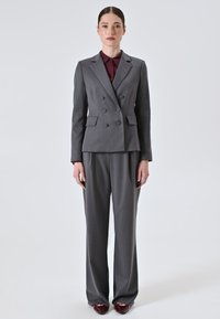 Giacca di suit grigia doppiopetto con due tasche frontali, abbinata a pantaloni grigi coordinati e una camicia bordeaux con bottoni sottostante.