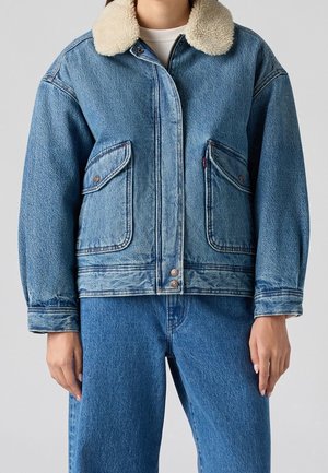 Personne portant une veste en denim bleu avec col sherpa beige et jean denim bleu assorti sur fond gris uni.