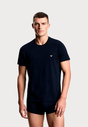 T-shirt di cotone blu navy con scollo rotondo e maniche corte, con un piccolo logo sul petto a sinistra. Indossata con pantaloncini abbinati.
