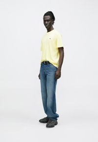 Maglietta gialla a maniche corte, vestibilità comoda, abbinata a jeans casual blu. Indossata con sneakers scure e acconciatura intrecciata.