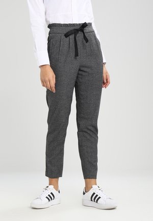 Personne portant un pantalon gris fuselé avec un cordon noir et des baskets blanches avec des rayures noires, se tenant devant un fond uni.