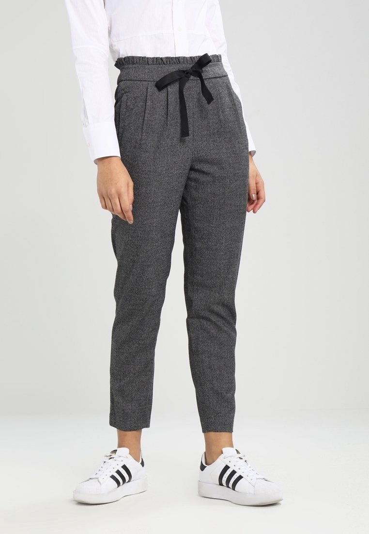 Personne portant un pantalon gris fuselé avec un cordon noir et des baskets blanches avec des rayures noires, se tenant devant un fond uni.