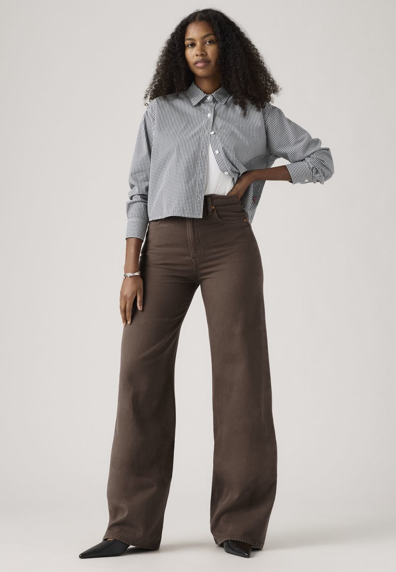 Femme aux cheveux bouclés portant une chemise cropée à carreaux noir et blanc sur un haut blanc, un pantalon large marron et des chaussures noires pointues.