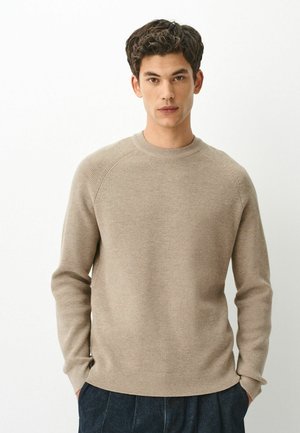 Young man with short curly hair wearing a beige knit sweater and dark pants, standing with hands in pockets against a white background.