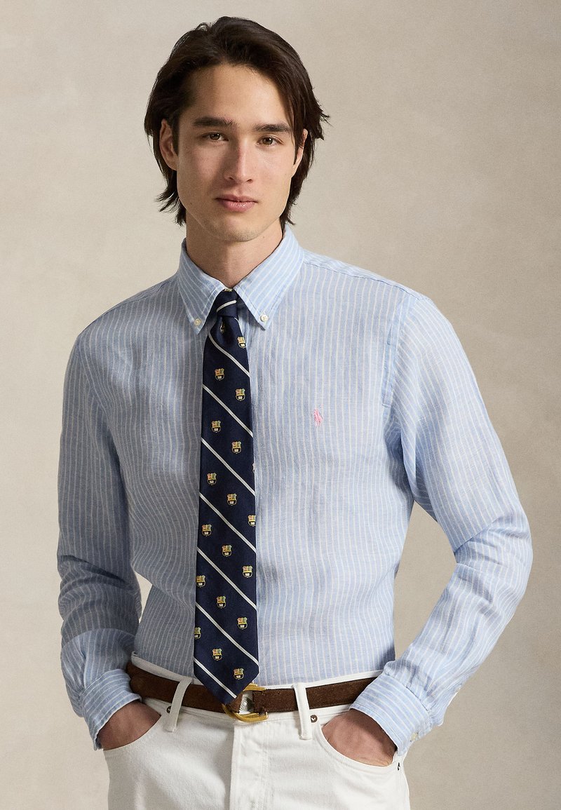 Noormees, kes kannab helesinist triibulist nööpidega särki, tumesinist mustrilist lipsu, valgeid pükse ja pruuni vööd, seisab käed taskutes.