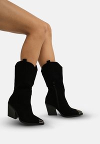 Botas de tobillo de ante negro con punta afilada, puntera metálica y tacón apilado, con cremallera lateral y costuras decorativas.