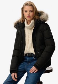 Sort pufferjakke med en pelsforing i hætten, der har lynlås og trykknaplukning, båret over en cremet ribbet sweater, kombineret med blå jeans.