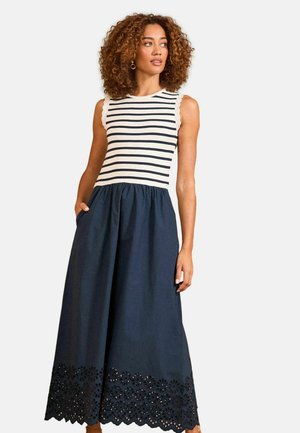 Žena s kučeravými vlasmi, ktorá nosí šaty bez rukávov s bielym a čiernym pruhovaným topom a námornícky modrú sukňu s kvetinovým výrezovým lemom, ruky vo vreckách.