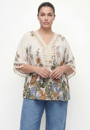 Vrouw met donker haar naar achteren gedragen draagt een witte bloemenblouse met kwastjes aan mouwen en halslijn, gecombineerd met blauwe spijkerbroek, staand tegen een witte achtergrond.