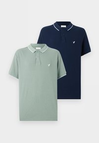 Δύο διπλωμένα κοντομάνικα πόλο, ένα ανοιχτοπράσινο και ένα σκούρο μπλε, το καθένα με λευκή διακόσμηση στον γιακά και στα μανίκια και μικρό λογότυπο με πουλί στο στήθος.