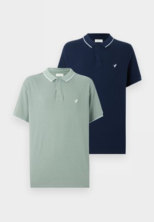 Homme debout portant un polo vert clair, un jean bleu ample, des chaussures noires et une montre-bracelet sur un fond blanc uni.