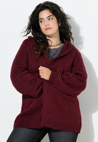 Donna con lunghi capelli ricci che indossa un cardigan spesso color bordeaux sopra una maglietta grigia, tersendo il cardigan chiuso, in piedi contro uno sfondo bianco.