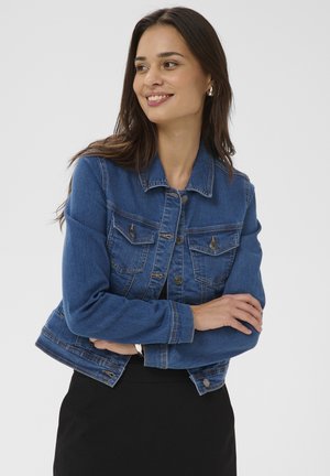Jeune femme aux longs cheveux bruns portant une veste en jean bleue et une jupe noire, les bras croisés, souriante et regardant sur le côté.