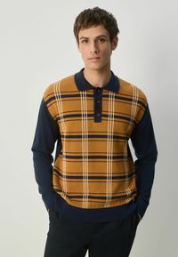 Strik polo sweater kombinerer orange-brun med marineblå ærmer. Har vandrette striber og tre knapper ved kraven. Blød tekstur.