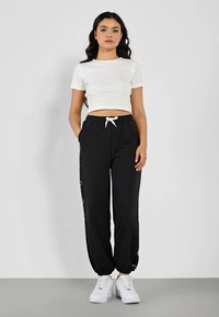 Schwarze Jogginghosen mit elastischen Bündchen und weißem Kordelzug, mit seitlichen Schlaufenakzenten. Kombiniert mit einem eng anliegenden weißen Crop-Top und weißen Sneakern.