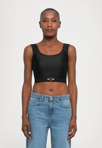 Woman wearing a black crop top with gold clasp detail and blue high-waisted jeans, standing against a plain white background.