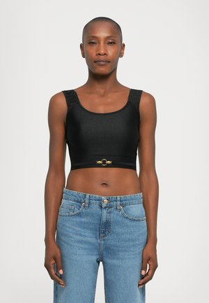 Woman wearing a black crop top with gold clasp detail and blue high-waisted jeans, standing against a plain white background.