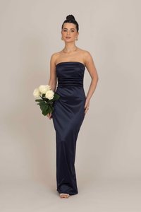 Απαλή ναυτική σατινέ φόρεμα χωρίς τιράντες με εφαρμοστό κορσάζ, λεπτομέρειες με ραβδώσεις και ρέουσα φούστα. Το μοντέλο κρατάει μια ανθοδέσμη με λευκά τριαντάφυλλα.