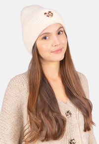 Bonnet en tricot côtelé crème avec un patch en forme de cœur en imprimé léopard. Le modèle porte un cardigan en tricot beige avec de gros boutons.
