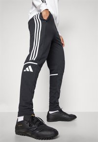 Black athletic pants with white stripes along the sides, featuring elastic waist and pockets. Worn with black sports shoes.