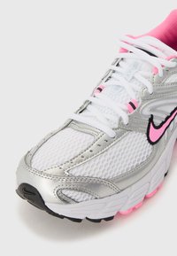 Chaussure de sport blanche avec tige en maille, accents argentés, détails roses, semelle extérieure noire et lacets blancs. Logo notable sur le côté.