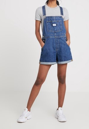Personne portant une salopette courte en denim bleu, un T-shirt gris clair et des baskets blanches, debout avec les mains dans les poches et les jambes écartées.