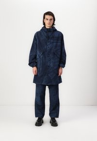 Jūras zila ūdensnecaurlaidīga jaka ar faktūru, kapuci un priekšējiem pogām, kas savienota ar atbilstošām brīvā stila biksēm un melnām kurpēm.