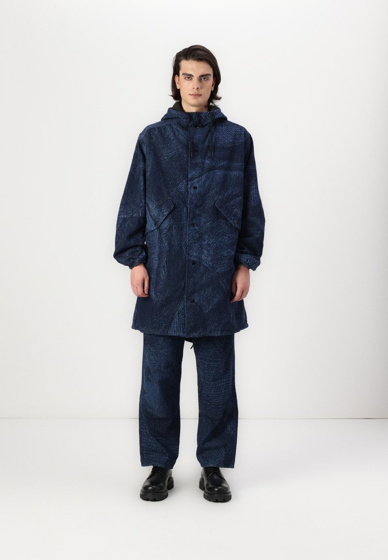 Jūras zila ūdensnecaurlaidīga jaka ar faktūru, kapuci un priekšējiem pogām, kas savienota ar atbilstošām brīvā stila biksēm un melnām kurpēm.