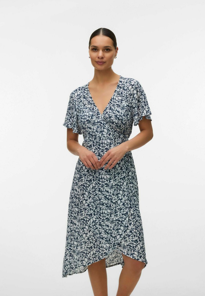 Femme portant une robe portefeuille mi-longue à fleurs bleu marine et blanche avec des manches courtes évasées, debout devant un fond blanc uni.