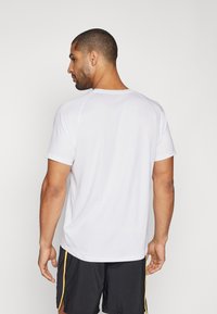 Camiseta atlética blanca de manga corta con cuello redondo, textura suave y costuras mínimas, combinada con pantalones cortos negros con acentos amarillos en los laterales.