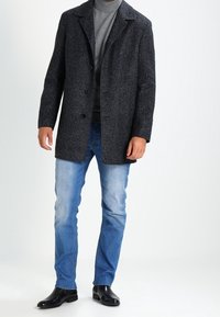 Abrigo de tweed gris oscuro con un cuello estructurado, tres botones y corte recto, combinado con jeans azul claro y zapatos de cuero negros.