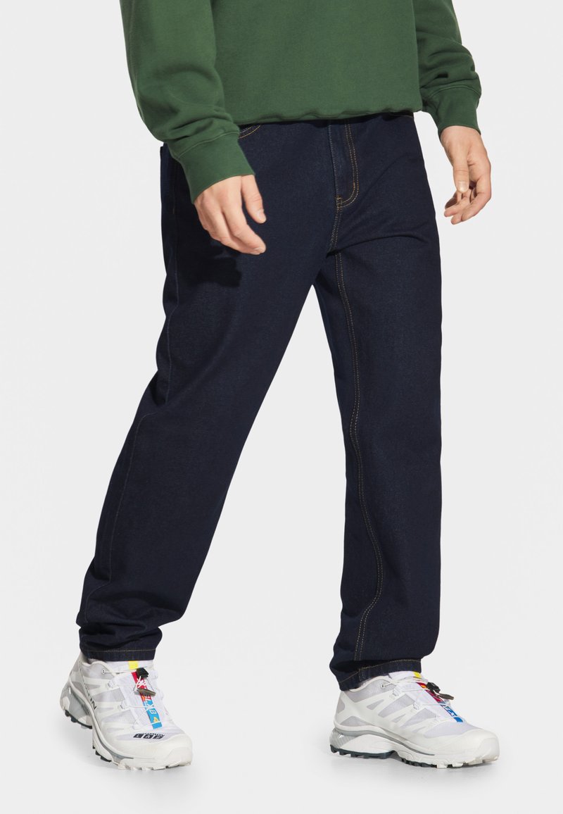 Jasnoniebieskie dżinsy z widocznymi kontrastowymi szwami. Luźny krój i wyraźne kieszenie; noszone z białymi trampkami z kolorowymi akcentami.