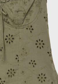 Tissu vert olive avec des motifs floraux brodés et des détails découpés, avec une couture froncée et nouée dans le coin supérieur gauche.