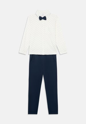 Camicia bianca a maniche lunghe con piccole pois nere e cravatta a farfalla blu navy abbinata a pantaloni blu navy con tasche laterali.