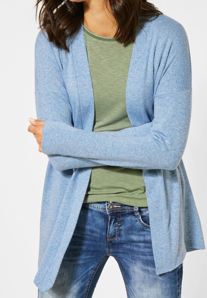 Femme portant un cardigan bleu clair ouvert sur un haut vert olive et un jean bleu, les bras croisés à la taille.