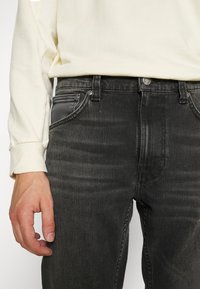 Černé džíny s úzkým střihem, které mají tradiční design pěti kapes a strukturovanou tkaninu. Zdobené černou koženou kapsou.