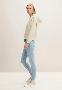 Beige huvtröja, ljusblå skinny jeans och ljusbeige hög sneakers. Modellen står på sidan mot en enfärgad bakgrund.