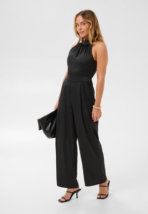 Femme portant une combinaison noire sans manches à col halter avec des jambes larges, tenant une pochette noire et portant des sandales à talons noires et un bracelet.