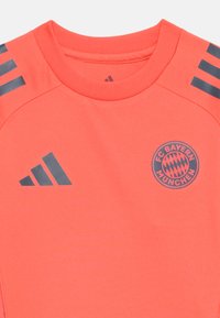 Camisa desportiva em coral brilhante com decote redondo, três riscas cinza-escuras em cada ombro e um logótipo circular do FC Bayern München.
