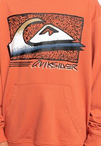 Sudadera naranja con un logotipo rectangular de estilo retro que presenta una ola y una montaña estilizadas, con el texto "QUIKSILVER" debajo.