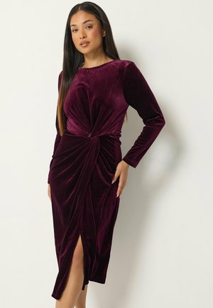 Femme portant une robe en velours violet profond à manches longues, avec un détail de nœud torsadé à la taille et une fente à l'avant, debout devant un fond uni.