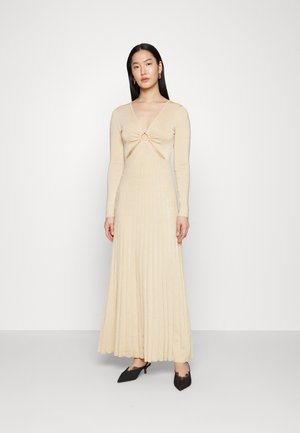 Γυναίκα που στέκεται σε μια μακριά μπεζ πλισέ φόρεμα με βαθύ V ντεκολτέ και κυκλική κοπή, φορώντας μαύρες αιχμηρές γόβες slingback.