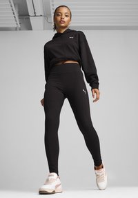 Czarna krótka bluza z kapturem i legginsy z wysokim stanem wykonane z elastycznego materiału. Białe sneakersy z beżowymi akcentami. Logo umieszczone na odzieży i obuwiu.